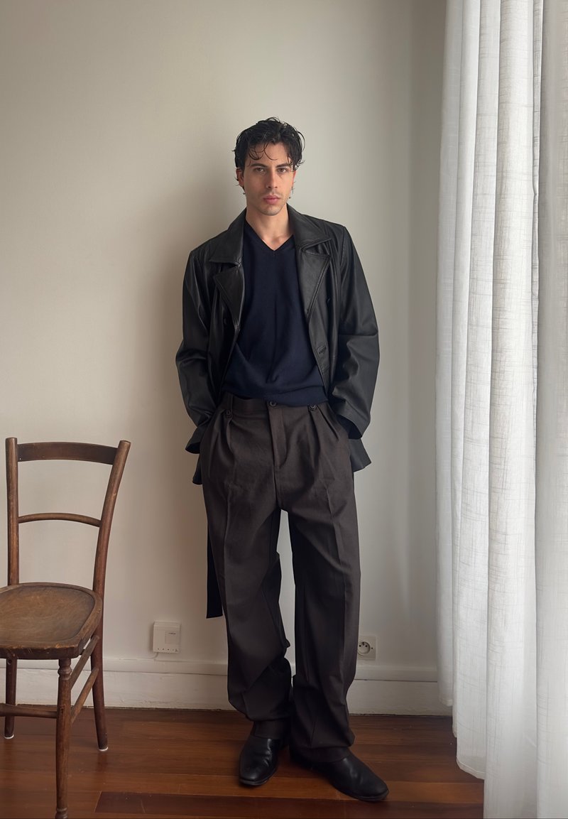 Young man with black leather jacket, black shirt, and loose dark trousers stands next to a wooden chair and white curtain in a minimalist room.