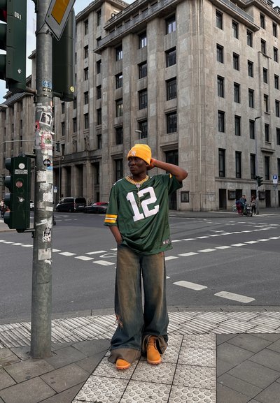Jersey verde con acentos amarillos y el número 12, pantalones de denim anchos desgastados, zapatillas naranjas y un gorro amarillo, de pie junto a un semáforo.