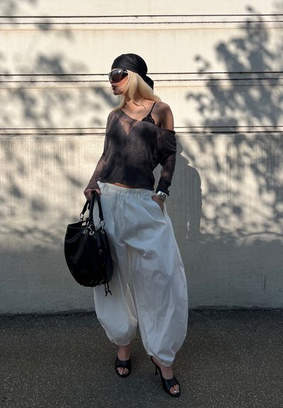 Mujer con una blusa negra transparente, pantalones balón blancos, pañuelo negro en la cabeza, gafas de sol y sandalias con tacón, sosteniendo un bolso negro frente a una pared clara.