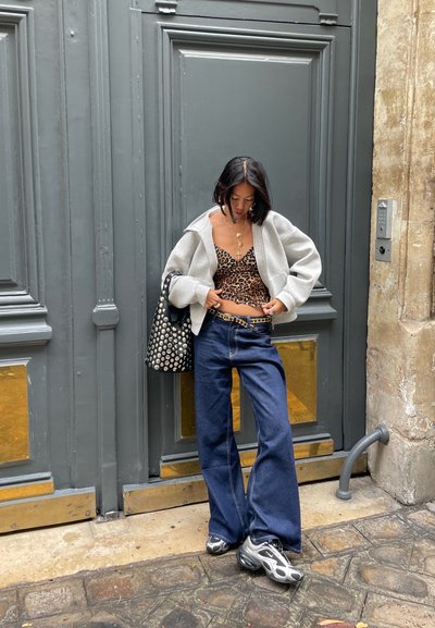 Mujer con una blusa de estampado de leopardo, chaqueta gris, jeans de pierna ancha y zapatillas, de pie en una calle de adoquines frente a una gran puerta gris.