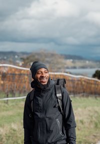 Lächelnder Mann mit schwarzer Mütze, Jacke und Rucksack, der in einem grünen Feld steht, mit einem Weinberg und bewölktem Himmel im Hintergrund.