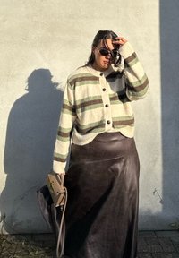 Krémový kardigán s zelenými a hnedými pruhmi, predné zapínanie na gombíky, nosený cez tmavohnedú koženú sukňu. Drží malú béžovú kabelku.