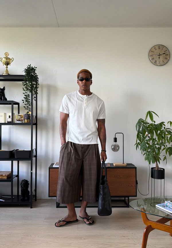 Man die een zonnebril draagt, wit overhemd, bruine wijde broek en slippers, een zwarte tas vasthoudend in een moderne kamer met planten en meubels.