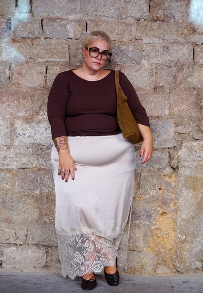 Top marrón de manga larga, falda crema con bordado de encaje y zapatos planos negros. Combinado con un bolso de hombro marrón. Pared de concreto en el fondo.