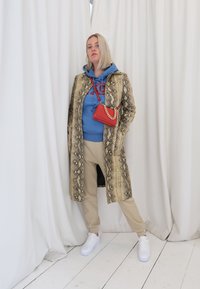 Abrigo largo de serpiente dorado, sudadera azul con logo rojo, pantalones beige y zapatillas blancas. Pequeño bolso rojo con detalles de cadena. Fondo blanco.