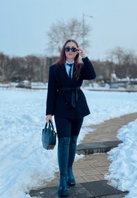 Blazer noir sur mesure avec ceinture à la taille, chemise blanche, cravate noire, leggings noirs brillants et bottines à plateforme épaisses. Porte un sac à main texturé.