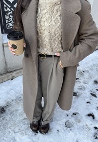 Άτομο στέκεται στο χιόνι φορώντας καφέ παλτό, μπεζ πουλόβερ με πλεκτό σχέδιο καλώδιο, ταουπέ παντελόνια με μαύρη ζώνη, καφέ μπότες, κρατώντας ένα ποτήρι καφέ για το δρόμο.