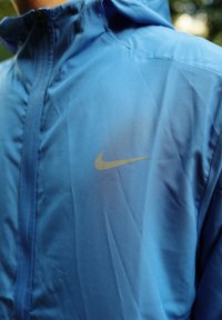 Blå Nike vindjakke med en glat tekstur, fuld lynlås og et diskret guld-logo på brystet. Letvægtig, velegnet til udendørs aktiviteter.