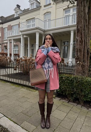 Joven con abrigo rosa, falda a cuadros, bufanda de cuadros y botas marrones, de pie en la acera frente a una casa de estilo victoriano con balcón blanco.