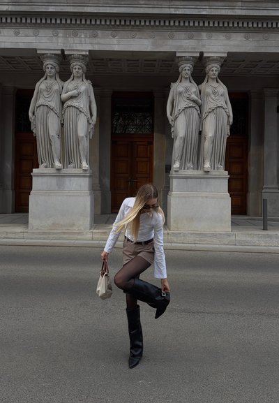 Mujer con camisa blanca y falda marrón ajusta bota negra hasta la rodilla en la calle frente a un edificio clásico con cuatro estatuas femeninas.