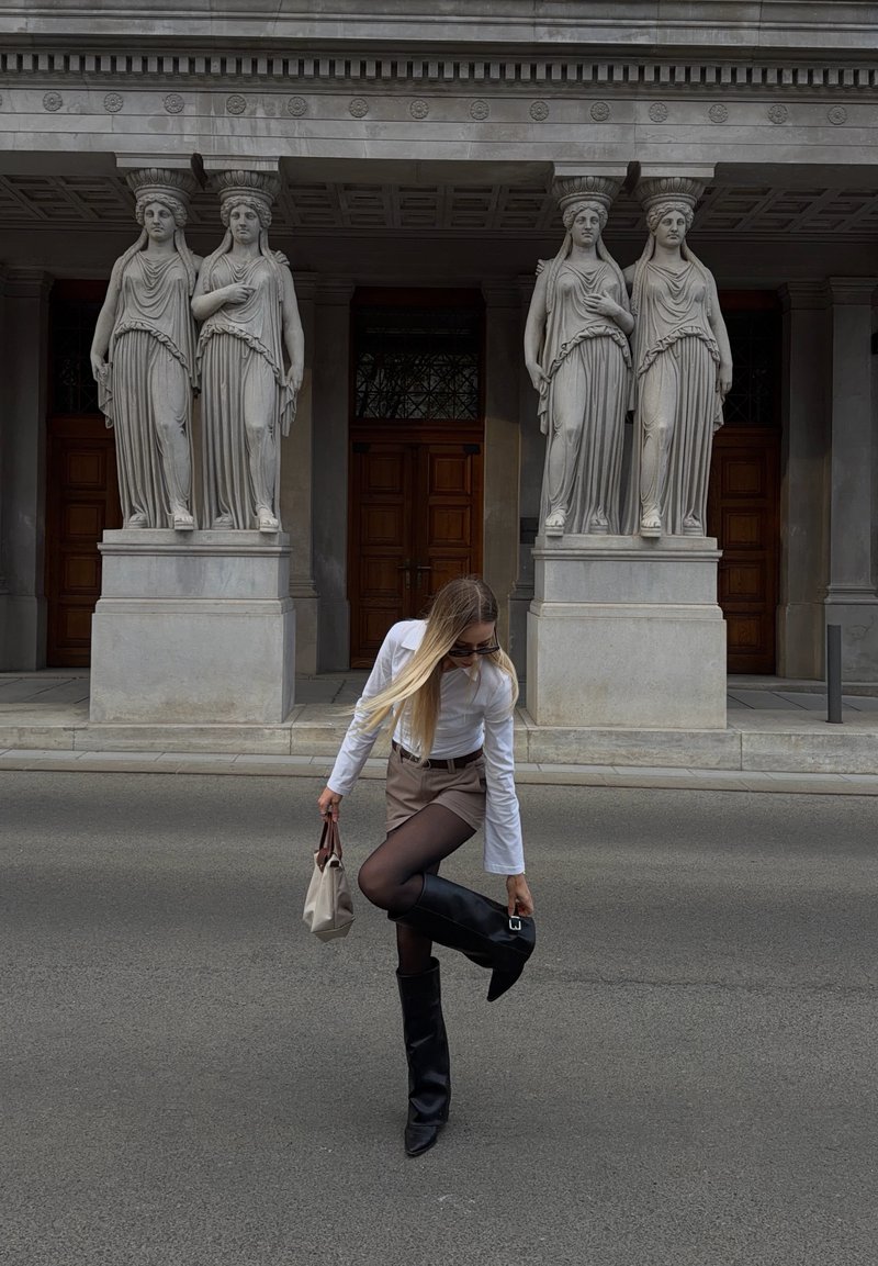 Frau in weissem Hemd und braunem Rock richtet schwarzen kniehohen Stiefel auf der Strasse vor einem klassischen Gebäude mit vier weiblichen Statuen.
