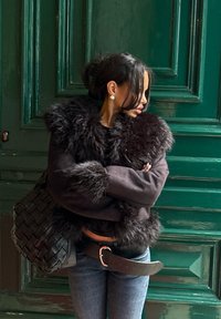 Fekete műszőrme dzseki széles gallérral, sötét farmernadrággal és egy nagy, szőtt fekete táskával párosítva. A háttér zöld fából készült panelekből áll.