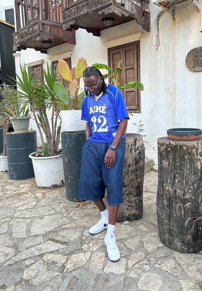 Jersey de malla azul con "AIMÉ 32" impreso, combinado con pantalones cortos de mezclilla oscura. Zapatillas Nike blancas y calcetines hasta el tobillo. Elementos naturales de fondo.