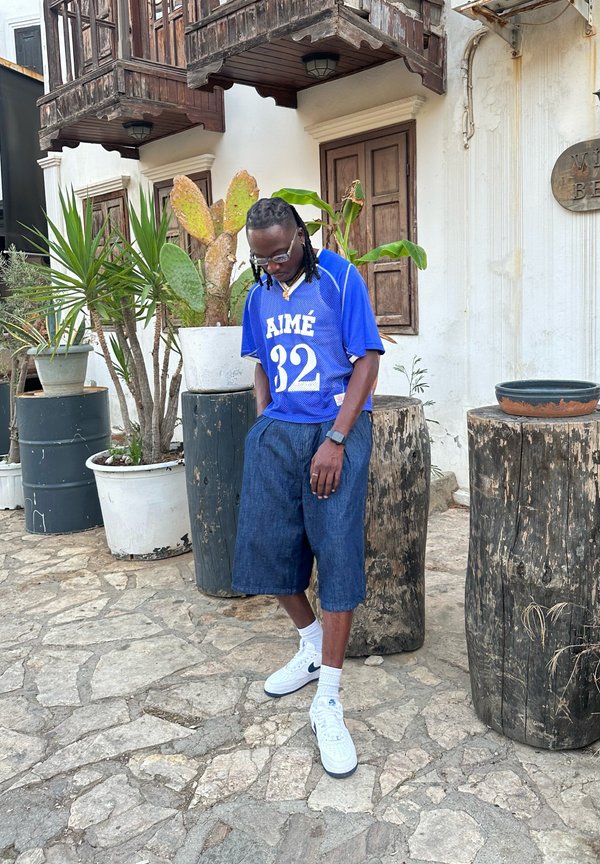 Kék hálós mez, amelyen "AIMÉ 32" van nyomtatva, sötét farmer rövidnadrággal párosítva. Fehér Nike sportcipő és bokazokni. Természetes háttérelemek.