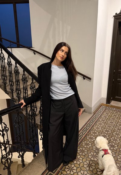 Abrigo largo negro sobre una blusa gris claro, combinado con pantalones anchos negros. Azulejos decorativos con patrón en el suelo y un perro blanco cerca.