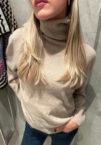 Μπεζ πουλόβερ με ψηλό γιακά σε μαλακό υφαντό πλέγμα, συνδυασμένο με σκούρα τζιν. Μακριά ξανθά μαλλιά πέφτουν στους ώμους.