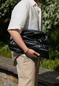 Μαύρη δερμάτινη τσάντα που κρατιέται ενάντια σε ένα ανοιχτόχρωμο πουκάμισο. Διαθέτει απαλή υφή και ρυθμιζόμενο λουρί με διακοσμητική αγκράφα.