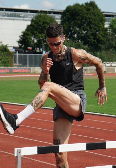 Atleta con un top negro sin mangas y pantalones cortos grises saltando vallas en una pista. Tatuajes visibles en brazos y piernas; lleva gafas de sol y zapatillas deportivas.
