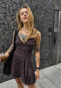 Μαύρο φόρεμα με κόκκινο λουλουδάτο σχέδιο, V-λαιμόκοψη, εφαρμοστό μπούστο, φαρδιά φούστα. Άτομο που κρατάει ένα κρύο ποτό, φορώντας στρωτές αλυσίδες και τατουάζ.