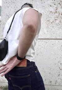 Weißes ärmelloses Shirt kombiniert mit dunkelblauen Denim-Jeans. Schwarze Schultertasche, sichtbares Markenetikett an den Jeans. Hintergrund zeigt eine strukturierte Wand.