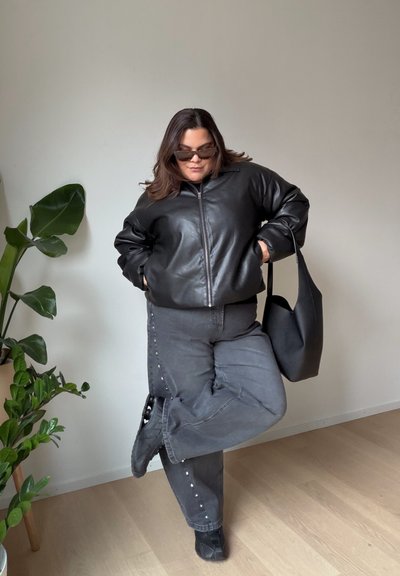 Chaqueta negra de cuero con cremallera y ajuste holgado, combinada con pantalones grises con remaches plateados. Los accesorios incluyen gafas de sol negras y bolso tote.