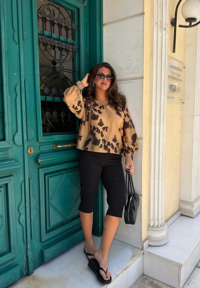 Mujer con blusa de estampado animal, pantalones capri negros, sandalias y gafas de sol, de pie junto a una puerta verde sosteniendo un bolso negro.