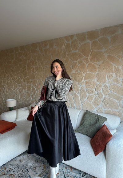 Mujer con falda midi negra, chaqueta de cuadros y botas blancas, de pie junto a un sofá blanco con cojines naranjas y verdes en una habitación con pared de piedra.