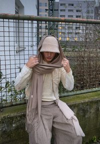 Μπεζ κασκόλ από μάλλινη ύφανση ριχτό στους ώμους, υφασμάτινη λευκή μπλούζα με υφή, ανοιχτό μπεζ καπέλο τύπου bucket και χαλαρές ταμπά παντελόνες. Κοσμήματα με πολλαπλούς δακτυλίους.