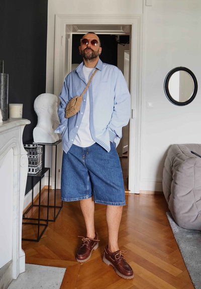 Shorts de mezclilla con un ajuste relajado, camisa de cuadros oversized en azul y blanco con botones, zapatos marrones de cordones y bolsa cruzada de tejido.