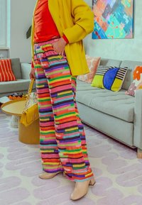 Bunte, gestreifte Hosen mit weitem Bein in leuchtendem Pink, Grün, Blau und Gelb. Kombiniert mit einem roten Oberteil und einem gelben Mantel, in einem gemütlichen Wohnzimmer.