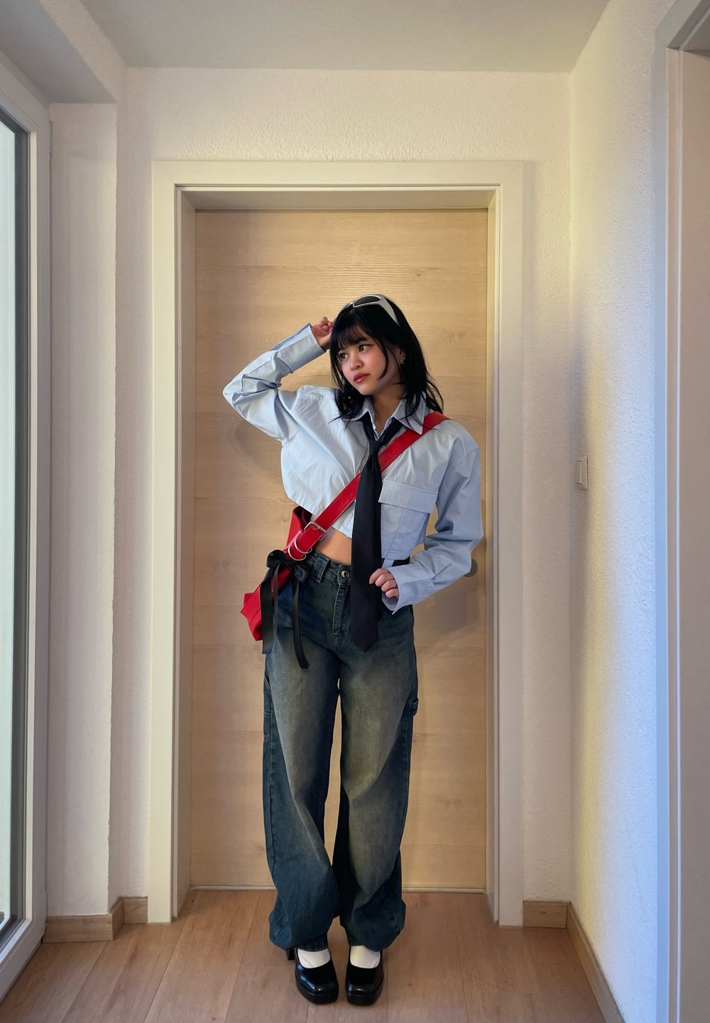 Camicia azzurra chiara tagliata con cravatta nera, borsa a tracolla rossa e jeans scuri larghi; indossa scarpe con plateau e calzini bianchi; ambiente interno.