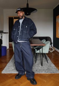 Donkerdenim jacket en brede spijkerbroek met contrasterende stiksels. Zwarte pet en schoenen. De kamer heeft een houten vloer en moderne meubels.