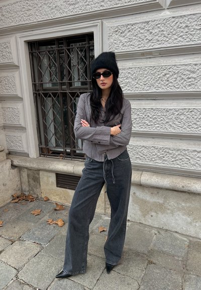 Camisa de botones a rayas con mangas anchas, combinada con jeans negros de cintura alta y botas negras puntiagudas, complementada con un gorro negro y gafas de sol.