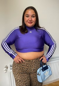 Vijoličasta skrajšana dolga majica z dolgimi rokavi in belimi črtami, v kombinaciji z hlačami z visokim pasom v leopardjem vzorcu. Drži svetlo modro torbico Adidas.