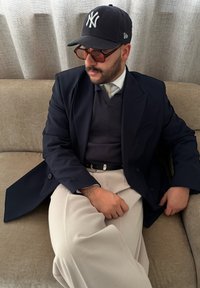 Σκούρο ναυτικό σακάκι πάνω από έναν γκρι πουλόβερ, κρεμ παντελόνι και μαύρο καπέλο μπέιζμπολ. Φοράει μεγάλα γυαλιά ηλίου και ασημένια βραχιόλι.