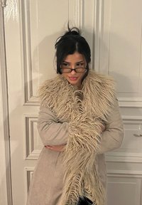 Manteau beige avec un long col en fausse fourrure. Le manteau est texturé et s'enroule autour du corps. Il présente un design simple et est associé à des lunettes.