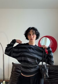 Haut à manches longues rayé noir et gris avec une taille froncée, assorti d'un sac à main noir. En arrière-plan, un canapé en cuir marron et un miroir circulaire rouge.