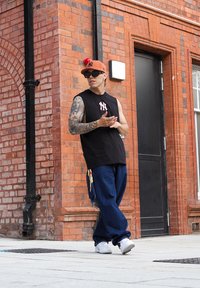 Črna majica brez rokavov, modri široki džins, bele teniske, oranžna kapa NY z rdečo pom-pom, sončna očala, tetovirano oko, stoječa ob rdeči opečni steni.