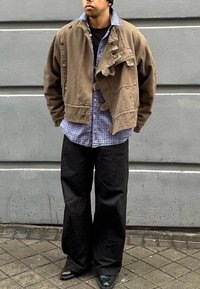 Barna pamut dzseki egyedi aszimmetrikus dizájnnal, nyomógombokkal és bolyhos gallérral, egy kék kockás ing felett és fekete széles nadrággal.