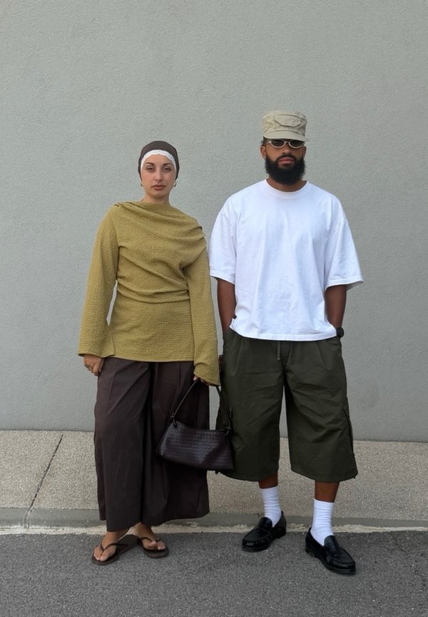 Une femme portant un foulard, un pull oversize, un pantalon large et des sandales se tient à côté d'un homme portant une casquette, des lunettes de soleil, un t-shirt blanc, un short long et des mocassins.