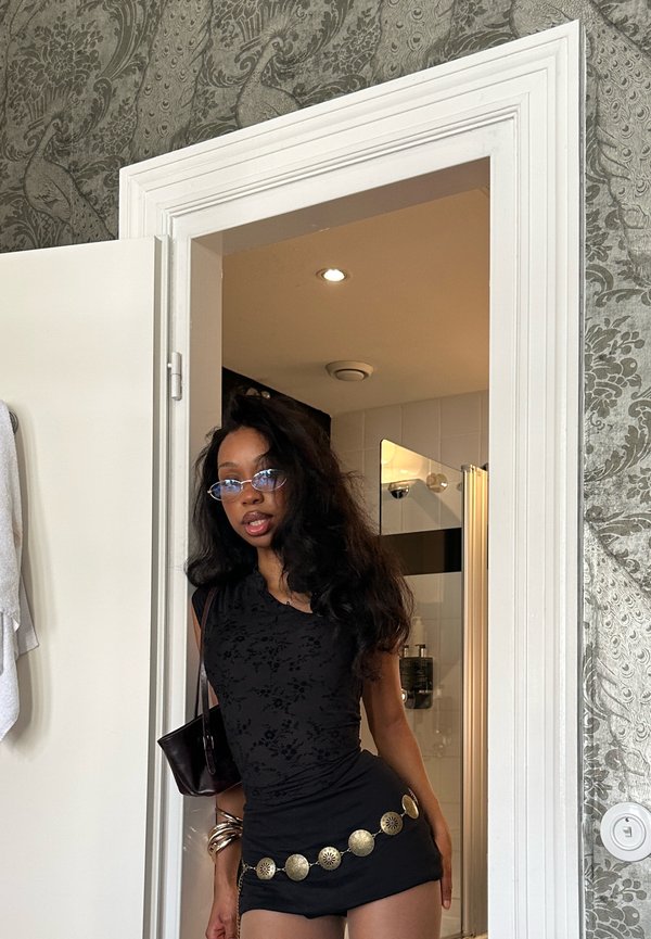 Młoda kobieta z długimi włosami, w okularach, czarnej bluzce i mini spódnicy z złotym paskiem, stojąca w drzwiach z tapetą w wzory.