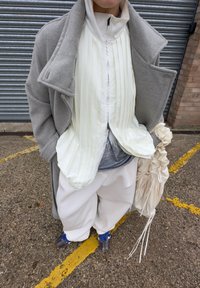 Manteau en laine gris clair sur une veste zippée blanche à rayures fines, associé à un pantalon blanc ample et des chaussures à motifs. Tient une pochette beige.