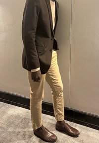 Personne debout à l'intérieur portant un blazer foncé, une chemise blanche, un pantalon beige, des mocassins marron et une bague à la main gauche.