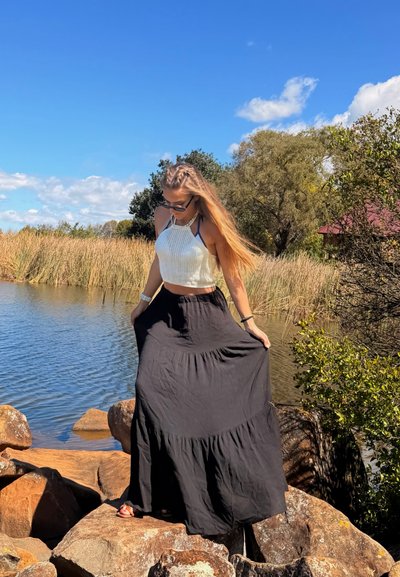 Joven con gafas de sol, top blanco de tirantes y falda larga negra, de pie sobre unas rocas junto a un cuerpo de agua con hierba alta y árboles.
