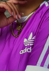 Maglietta Adidas viola con strisce bianche, con un logo ben visibile. Il tessuto è liscio, con una sottile texture verticale. Collane a strati dorate in vista.