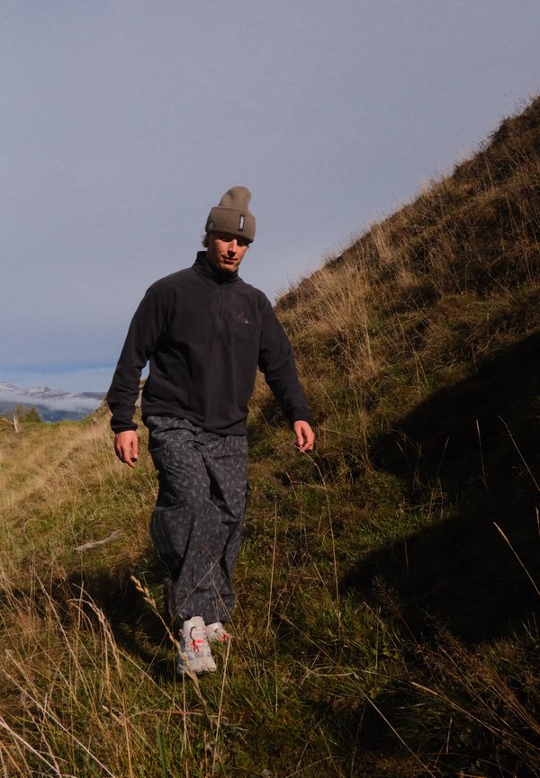 Un excursionista con gorro de lana, chaqueta oscura y pantalones estampados camina entre hierba alta por una ladera soleada.