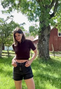 Kürzlich geschnittenes burgunderfarbenes Polohemd mit kurzen Ärmeln und gestrickter Textur, kombiniert mit hochgeschnittenen schwarzen Denim-Shorts. Hintergrund aus grünem Gras und Bäumen.