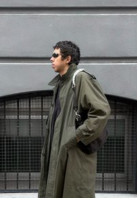 Jeune homme portant des lunettes de soleil noires et un long manteau vert olive, tenant un sac noir avec une bandoulière blanche, debout contre un mur gris.
