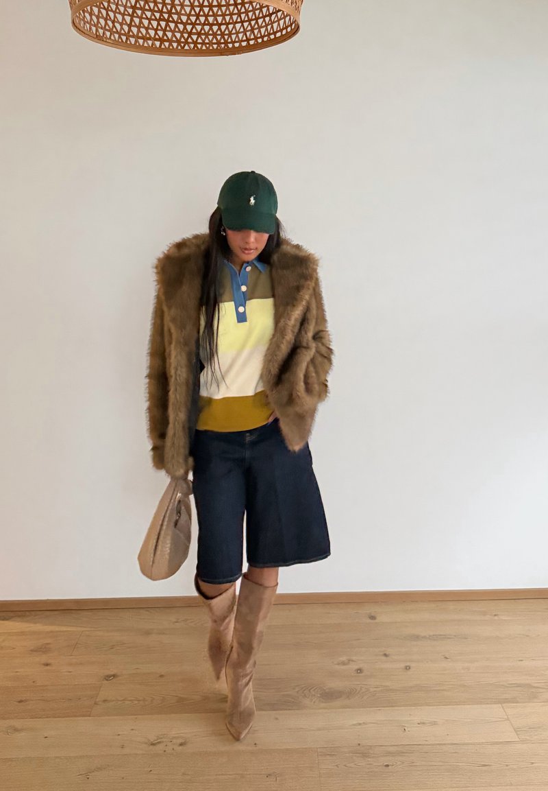 Donna che indossa una giacca di pelliccia, camicia a blocchi di colore, pantaloncini di jeans, stivali beige e berretto verde, con una borsa beige, in piedi al chiuso.