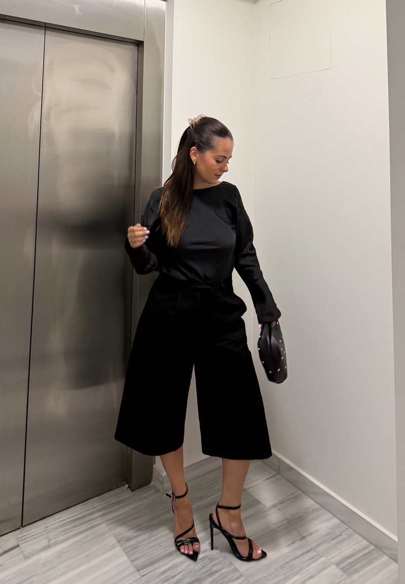 Woman in black long-sleeve top and wide-leg cropped pants wearing black high heels, holding a small handbag near an elevator.